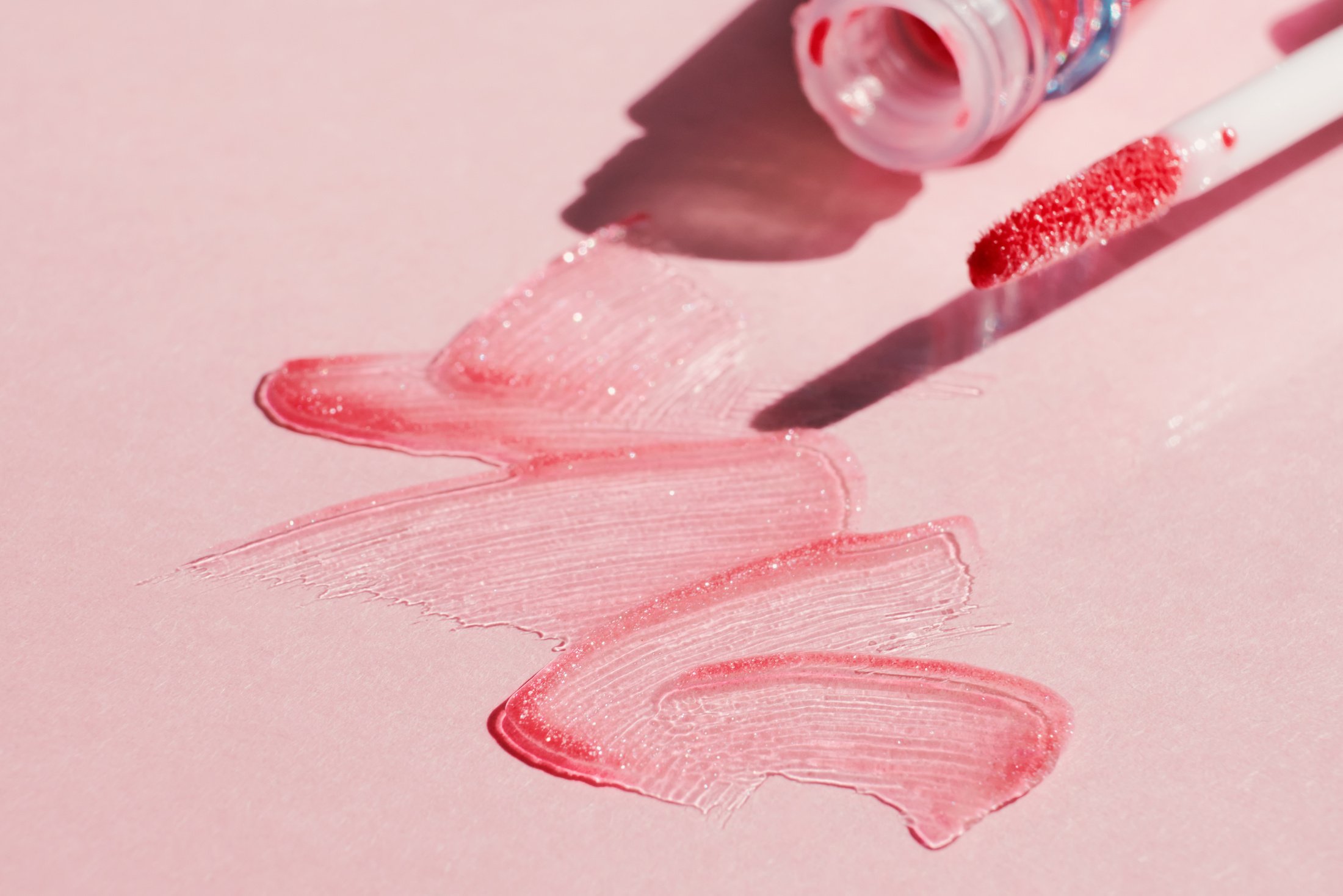 Smears of shining lip gloss and lip gloss brush on pink background, hard shadows